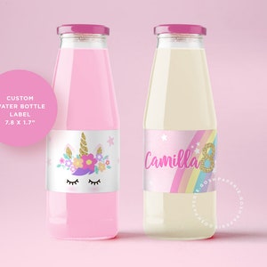 May include: Two clear glass water bottles with pink and white labels. The label on the left bottle features a unicorn with flowers and stars. The label on the right bottle features a rainbow, stars, and the text "Camilla 8".