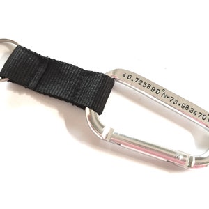 May include: Silver carabiner keychain with a black nylon strap and a key ring. The carabiner is engraved with the coordinates "40.725880&deg; N-73.983470&deg; W".