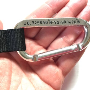 May include: A silver carabiner with a black strap attached. The carabiner has the coordinates "40.725890&deg; N-73.983470&deg; W" engraved on it.