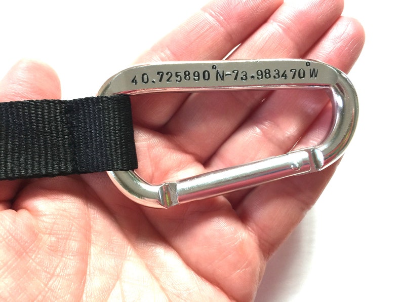 May include: A silver carabiner with a black strap attached. The carabiner has the coordinates "40.725890&deg; N-73.983470&deg; W" engraved on it.