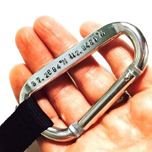 May include: A silver carabiner with a black strap attached. The carabiner has the coordinates "37.2694&deg; N 122.9481&deg; W" engraved on it.