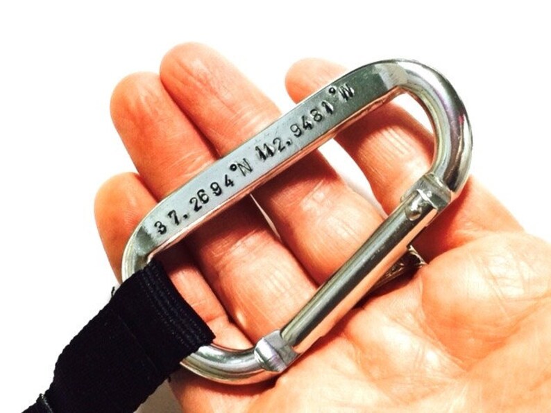 May include: A silver carabiner with a black strap attached. The carabiner has the coordinates "37.2694&deg; N 122.9481&deg; W" engraved on it.