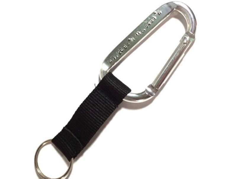 May include: A silver carabiner with a black strap and a key ring. The carabiner has the text "30.2694 N, 92.3431 W" engraved on it.