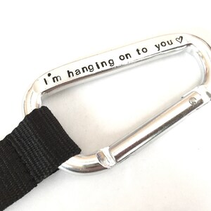 May include: A silver carabiner with a black strap attached. The carabiner has the words "I'm hanging on to you" engraved on it.