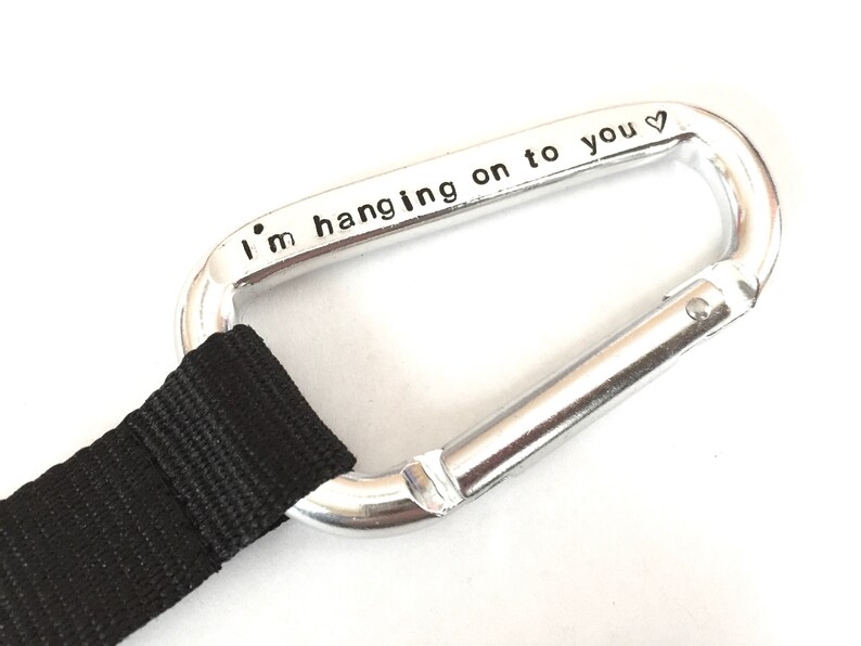 May include: A silver carabiner with a black strap attached. The carabiner has the words "I'm hanging on to you" engraved on it.