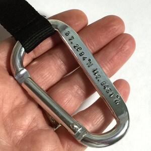 May include: A silver carabiner with a black strap attached. The carabiner has the coordinates "37.2694&deg; N 92.9481&deg; W" engraved on it.
