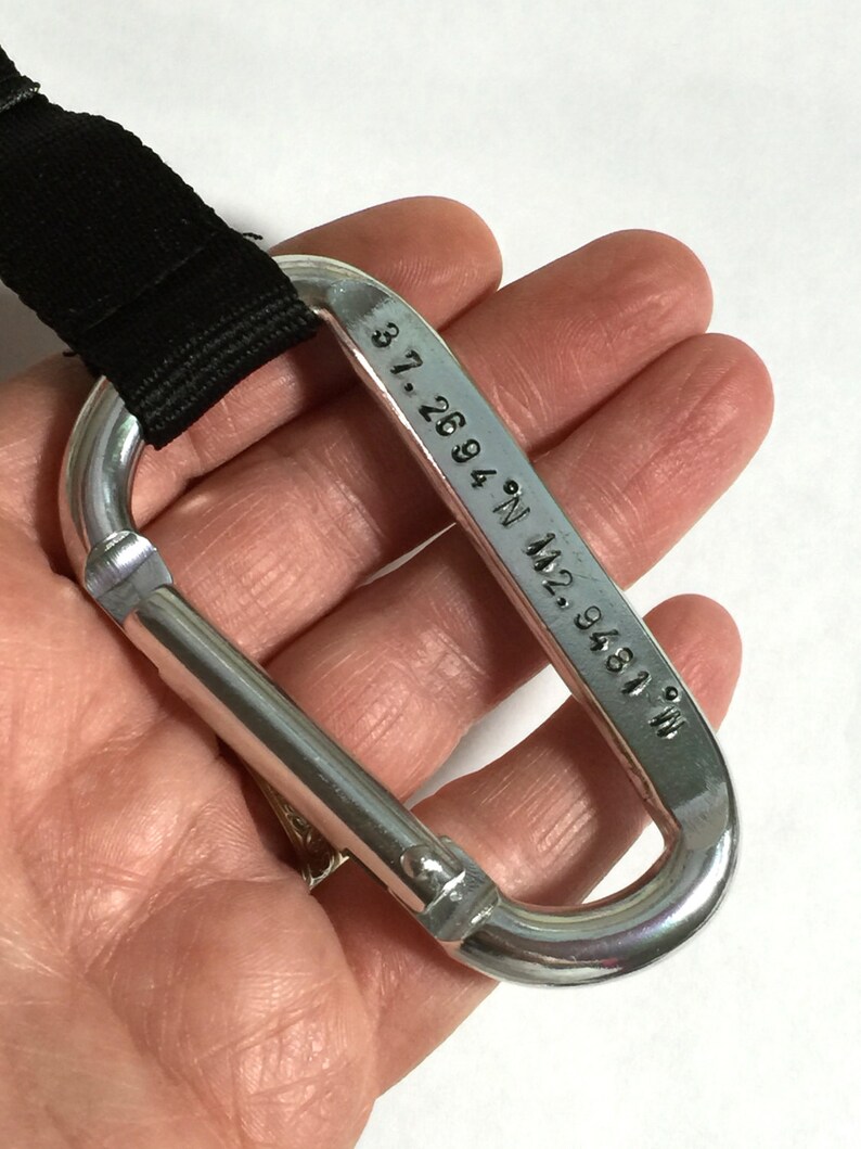 May include: A silver carabiner with a black strap attached. The carabiner has the coordinates "37.2694&deg; N 92.9481&deg; W" engraved on it.