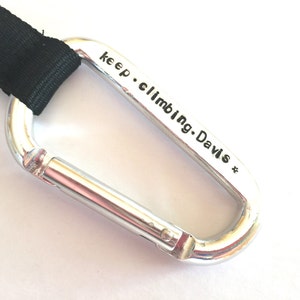 May include: Silver carabiner with a black strap attached. The carabiner has the text "keep climbing. Davis *" engraved on it.