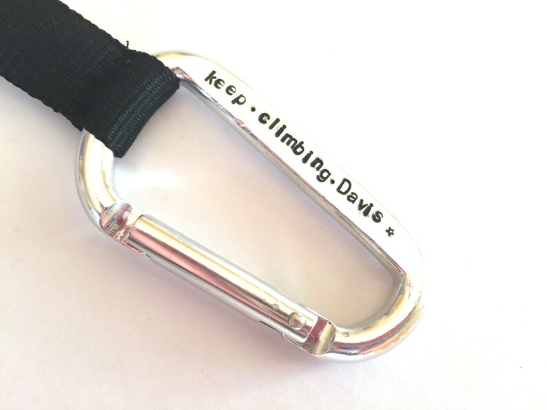 May include: Silver carabiner with a black strap attached. The carabiner has the text "keep climbing. Davis *" engraved on it.