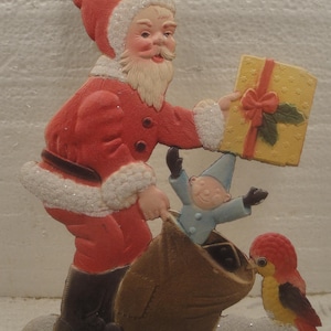 May include: A Christmas-themed decoration featuring Santa Claus in a red suit, holding a gift box and a sack. A small elf and a bird are also present. The words "A Merry Christmas" are at the bottom.