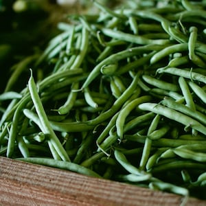 May include: A large pile of fresh, vibrant green beans packed tightly together. The slender, elongated pods feature a smooth, firm texture and a bright green color, filling a wooden container that is partially visible at the bottom of the frame.