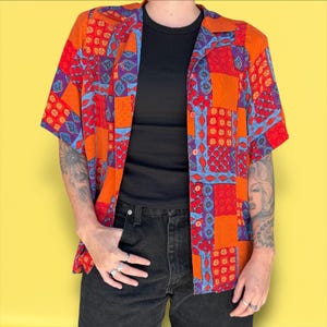 May include: Short-sleeved button-down shirt with a patchwork design in shades of red, orange, blue, and purple. The shirt has a collared neckline and is worn over a black top. The model is wearing black jeans.