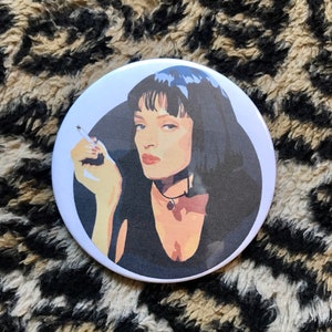 May include: A white button with a black and white illustration of a woman with dark hair and a cigarette in her hand. The woman is wearing a black top and a silver necklace.