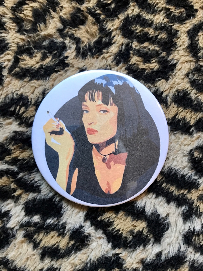 May include: A white button with a black and white illustration of a woman with dark hair and a cigarette in her hand. The woman is wearing a black top and a silver necklace.