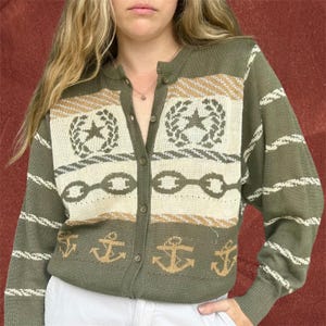 May include: Olive green cardigan sweater with a button-up front. The sweater features a cream-colored panel with star and wreath designs, chain links, and anchors. The sleeves have horizontal stripes. A classic nautical-themed knit.