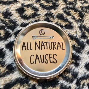 May include: A silver button with the text "ALL NATURAL CAUSES" in black lettering. The button has a crescent moon design on the top.