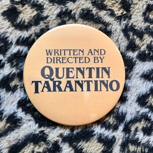 May include: A round, tan button with black text that reads "Written and Directed by Quentin Tarantino".