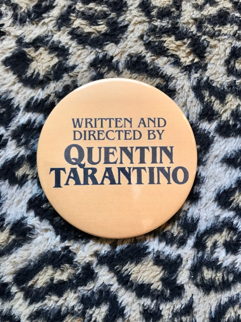 May include: A round, tan button with black text that reads "Written and Directed by Quentin Tarantino".
