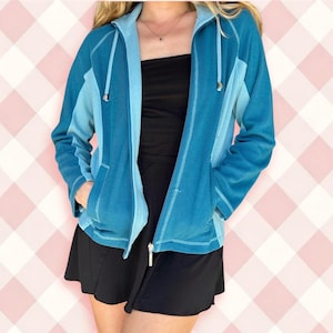 May include: A teal and light blue zip-up hoodie with a drawstring hood and front pockets. The jacket has a contrasting light blue panel on the shoulders and hood. The zipper is silver. The person is wearing a black skirt.