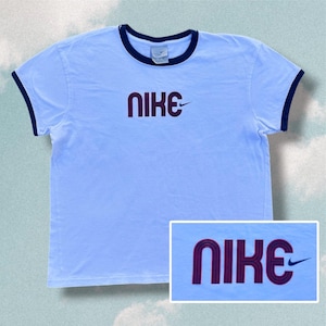 May include: A light blue short-sleeve t-shirt with a dark blue crew neckline and sleeve trim. The center of the chest features the word NIKE in a dark blue, multi-line graphic font with a small dark blue swoosh logo to the right of the text.