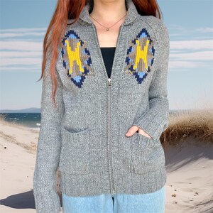 Könnte beinhalten: Graue Strickjacke mit Reißverschluss, zwei Vordertaschen und einem Kragen. Die Strickjacke hat ein geometrisches Design in Gelb, Blau und Braun auf der Brust. Sie hat einen silbernen Reißverschluss und ist aus dickem Strickmaterial gefertigt.