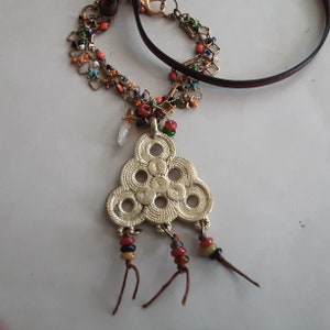 May include: A gold necklace with a triangular pendant featuring a repeating circle design. The pendant has three strands of brown leather with colorful beads hanging from it.