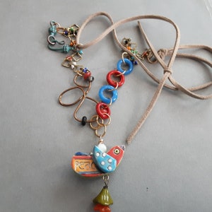 May include: A colorful necklace with a peace dove charm. The necklace features a suede cord, red and blue glass beads, and a gold chain with various charms. The peace dove charm is made of ceramic and has a blue and white design with the word "peace" written on it.