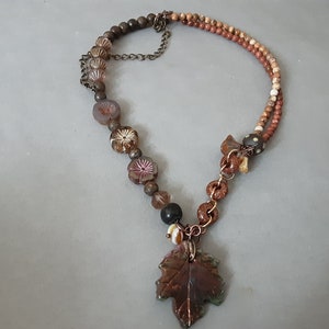 May include: A brown and gold necklace with a leaf pendant. The necklace is made of various beads, including brown, gold, and white beads. The leaf pendant is made of a brown material and has a green and brown design.
