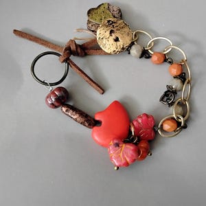 May include: A bracelet featuring a brown leather cord tie, a dark metal ring, and a gold-tone chain. The piece includes various beads in orange, red, and grey, along with a prominent red heart-shaped charm, leaf-shaped charms, and a textured gold-tone button.