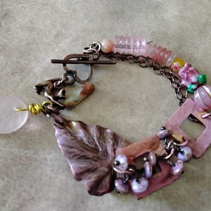 May include: A handcrafted bracelet featuring a variety of charms and beads. The bracelet includes a pink leaf-shaped charm, a copper-colored feather, and various pink and purple beads. The clasp is a toggle style, and the chain is a dark bronze color.
