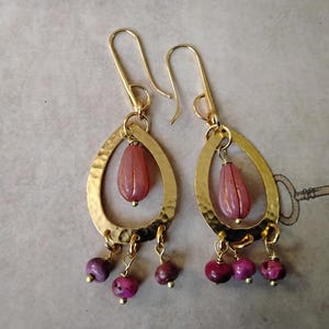 May include: Gold-tone earrings with a hammered teardrop shape. Each earring features a pink, teardrop-shaped bead suspended within the frame, and three smaller, round, purple beads hanging below. The earrings have a hook closure.