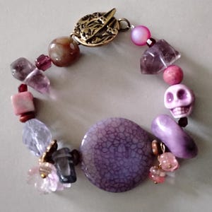 May include: A beaded bracelet featuring a variety of purple, pink, and brown beads. The bracelet includes a skull charm, a large cracked purple bead, and a gold-colored clasp. The beads are of various shapes and sizes, creating a unique and eye-catching accessory.