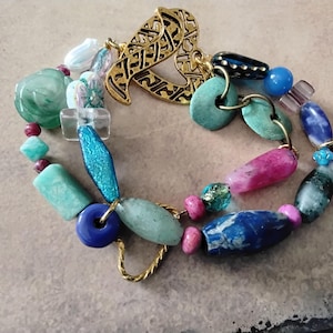 May include: A colorful beaded bracelet featuring a variety of shapes and colors. The bracelet includes turquoise, blue, pink, and green beads, with a gold-colored clasp. The design incorporates a mix of textures and sizes, creating a unique accessory.