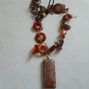 May include: A long necklace with a rectangular pendant made of brown and red stone. The necklace is made of brown chain, wooden beads, and red and brown glass beads.
