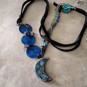 May include: Two handmade necklaces. One features a crescent moon pendant and blue disc accents. The other has a turquoise flower and beaded accents. Both have black suede cords and copper wire detailing. The jewelry is displayed on a neutral surface.
