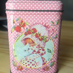 May include: A rectangular pink metal tin featuring a white polka dot pattern. The front displays a circular illustration of a character wearing a strawberry-patterned hat and dress, holding a white cat, surrounded by strawberry vines.