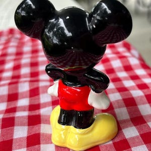 May include: A ceramic figurine of Mickey Mouse, featuring a glossy black head with large ears, a red shirt, black shorts, white gloves, and yellow shoes. The figurine is posed on a red and white checkered surface.