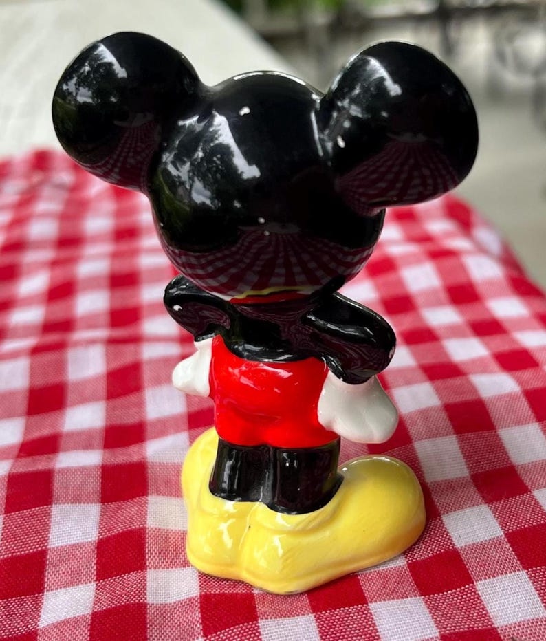 May include: A ceramic figurine of Mickey Mouse, featuring a glossy black head with large ears, a red shirt, black shorts, white gloves, and yellow shoes. The figurine is posed on a red and white checkered surface.