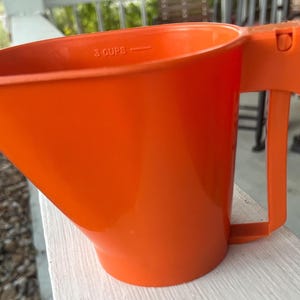 May include: Bright orange plastic measuring cup with a handle. The cup is marked with "3 CUPS" near the top. The handle is attached to the side of the cup and has a curved design for easy gripping. The cup is sitting on a white surface.