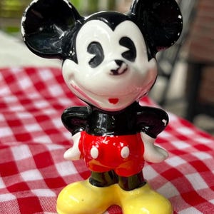 May include: A ceramic figurine of Mickey Mouse, featuring black ears, a white face, and a red shorts. The figure has yellow shoes and white gloves. The figurine is standing on a red and white checkered surface.
