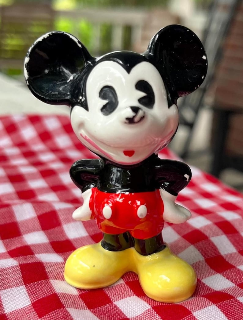May include: A ceramic figurine of Mickey Mouse, featuring black ears, a white face, and a red shorts. The figure has yellow shoes and white gloves. The figurine is standing on a red and white checkered surface.