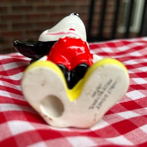 May include: A ceramic Mickey Mouse figurine in a reclining pose. The figure features a white head with black ears, a red torso, black shorts, and yellow shoes. The figurine is on a red and white checkered surface.