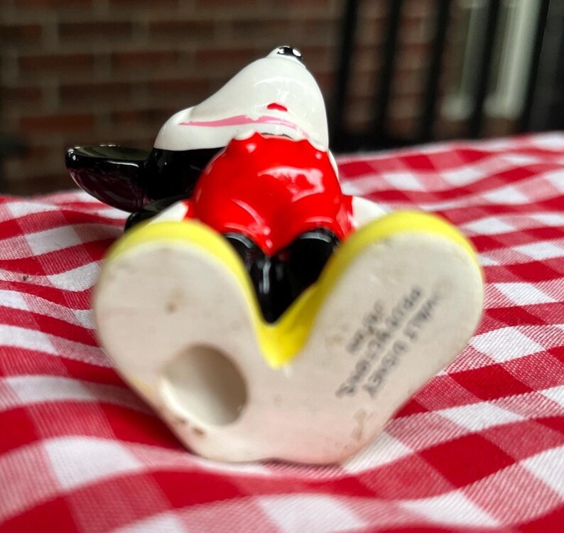 May include: A ceramic Mickey Mouse figurine in a reclining pose. The figure features a white head with black ears, a red torso, black shorts, and yellow shoes. The figurine is on a red and white checkered surface.
