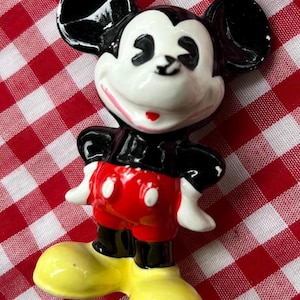 May include: A ceramic Mickey Mouse figurine, featuring a white face, black ears, and a red and black outfit. The figurine has yellow shoes and is posed with arms at its sides. The background is a red and white checkered pattern.