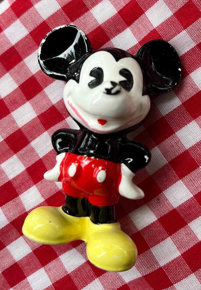 May include: A ceramic Mickey Mouse figurine, featuring a white face, black ears, and a red and black outfit. The figurine has yellow shoes and is posed with arms at its sides. The background is a red and white checkered pattern.