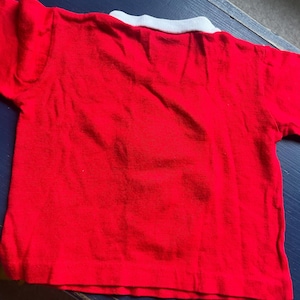 K&ouml;nnte beinhalten: Ein rotes Kurzarm-Shirt mit wei&szlig;em Kragen. Das Shirt besteht aus einem weichen Material und ist wahrscheinlich f&uuml;r ein Kind. Das Shirt liegt flach auf einer dunkelblauen Oberfl&auml;che.