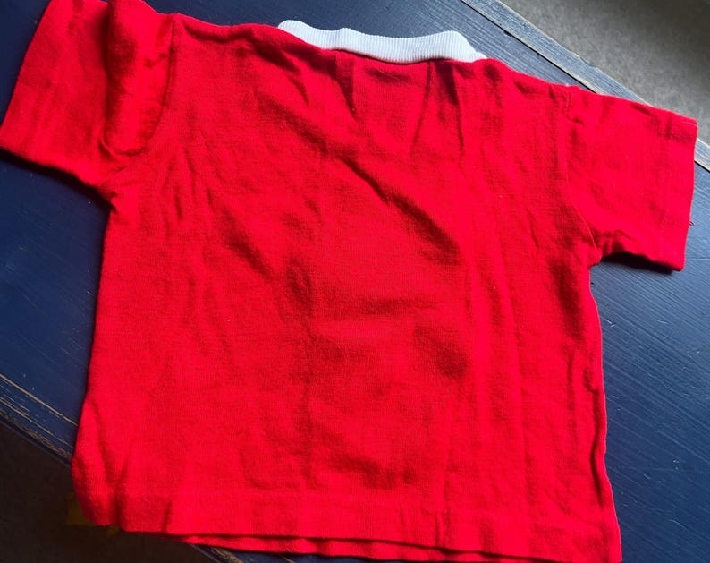 K&ouml;nnte beinhalten: Ein rotes Kurzarm-Shirt mit wei&szlig;em Kragen. Das Shirt besteht aus einem weichen Material und ist wahrscheinlich f&uuml;r ein Kind. Das Shirt liegt flach auf einer dunkelblauen Oberfl&auml;che.
