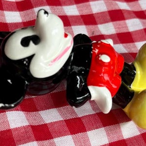 May include: A ceramic figurine of Mickey Mouse, featuring a white face with pink accents, black ears, a red torso, and yellow feet. The figure is lying on a red and white checkered surface.