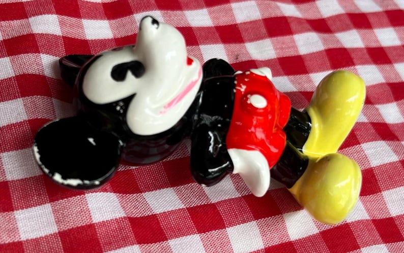 May include: A ceramic figurine of Mickey Mouse, featuring a white face with pink accents, black ears, a red torso, and yellow feet. The figure is lying on a red and white checkered surface.