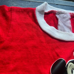 K&ouml;nnte beinhalten: Ein rotes T-Shirt mit wei&szlig;em Kragen und einer Mickey Mouse-Grafik. Das Shirt hat ein Etikett mit der Aufschrift "by P...". Das Shirt ist aus einem weichen Material gefertigt.